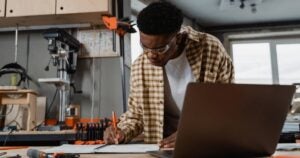A person wearing safety glasses and a plaid shirt working in a workshop, writing or drawing on a piece of paper with a pencil.