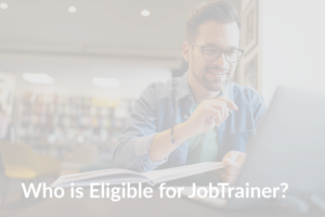 Main wearing glasses and a blue shirt sitting on a desk holding a book and looking at his laptop with the title 'Who is Eligible for JobTrainer' on the image.
