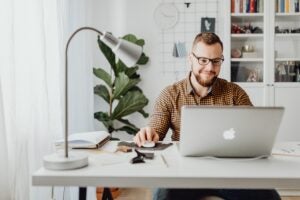A man with glasses and a beard working at a desk with a laptop in a home office. The background includes a bookshelf, a potted plant, and a desk lamp.